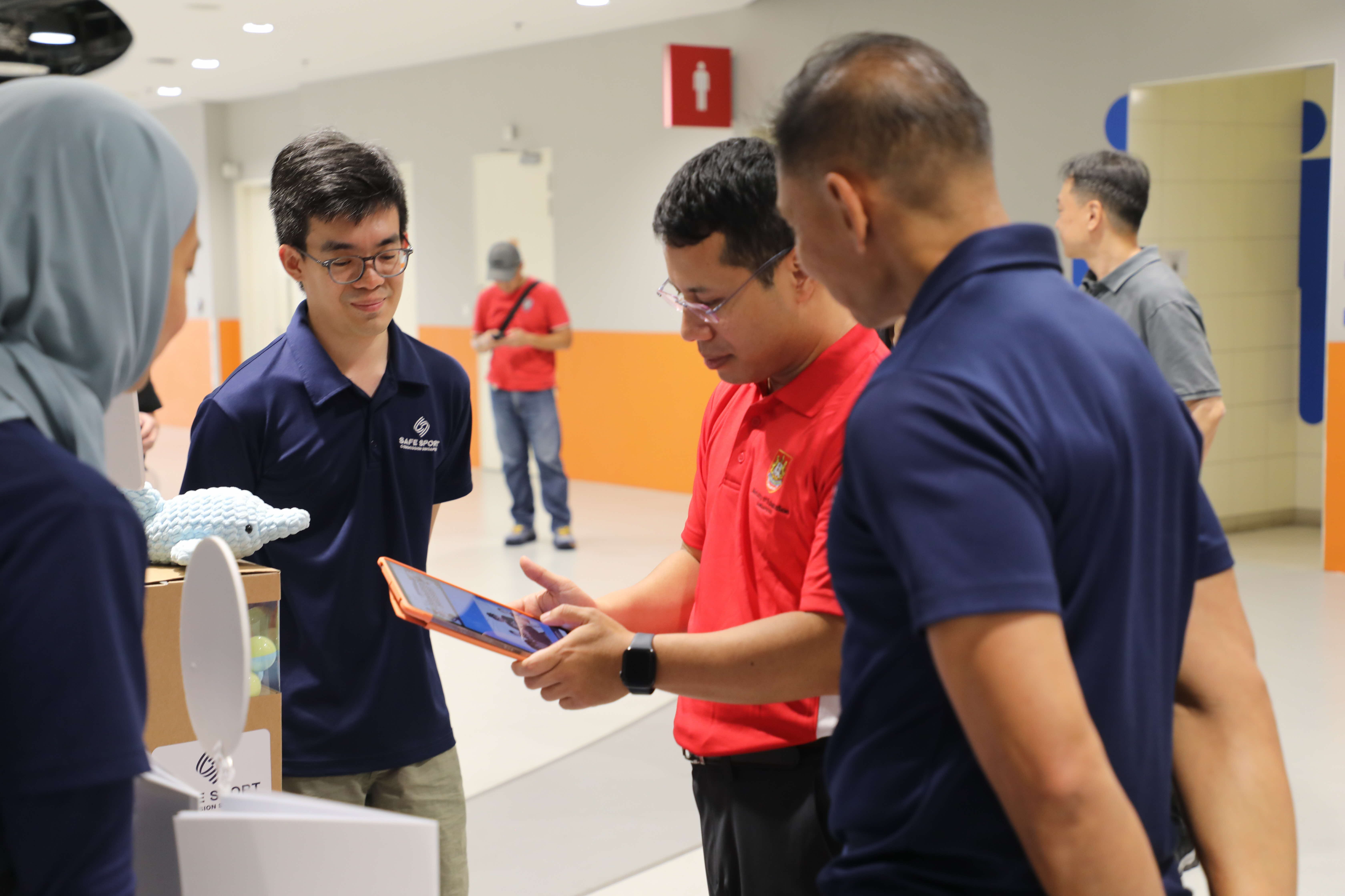 A group of people in a hallway, with one person in a red shirt showing something on a tablet to others. They appear engaged and focused.